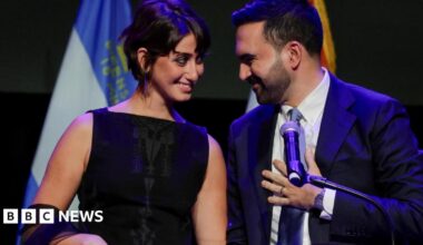 Zohran Mamdani styands behind a podium with his wife and is smiling at her. There are flags in the background and he is touching his tie.