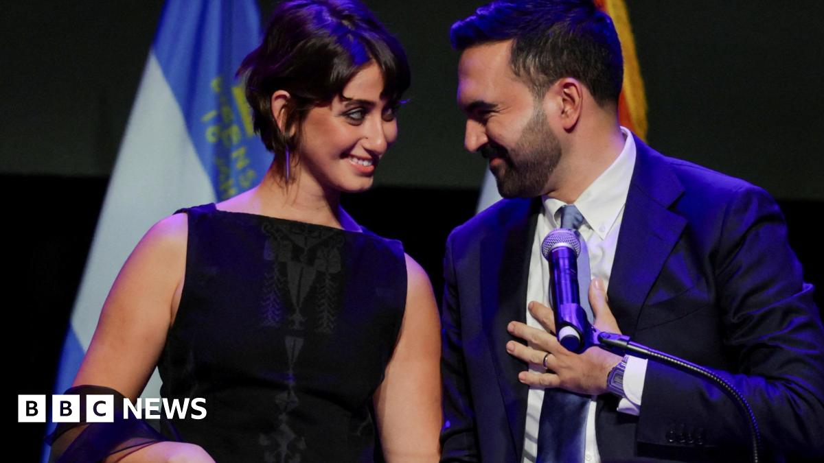 Zohran Mamdani styands behind a podium with his wife and is smiling at her. There are flags in the background and he is touching his tie.