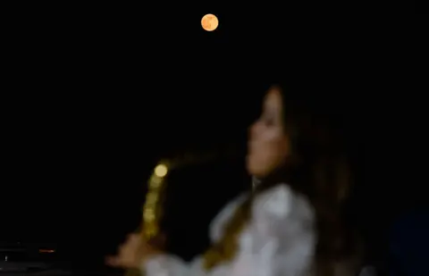 Ibraheem Abu Mustafa / Reuters The female musician can be seen in the foreground but out of focus, while the Moon shines very orange against a starless night sky in Doha on Wednesday.