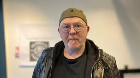 A man with a grey moustache and small goatee, wears round glasses in a thin frame. He is also wearing a black leather jacket with a black hoodie underneath, and is looking directly at the camera. 