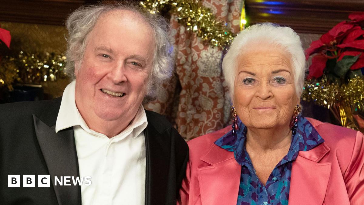 Nigel (Paul Bradley) with Pat (Pam St Clement) in character in the Queen Vic, in sitting and smiling in front of Christmas decorations