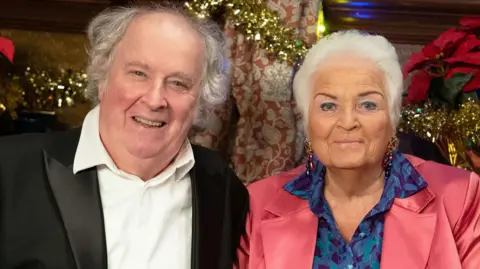 BBC Nigel (Paul Bradley) with Pat (Pam St Clement) in character in the Queen Vic, in sitting and smiling in front of Christmas decorations