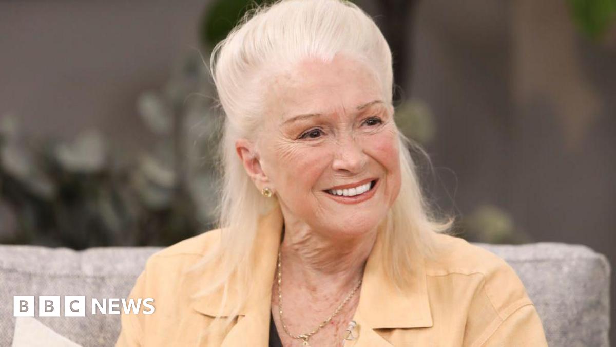 Actress Diane Ladd smiles while sitting on a couch. She is wearing a tan shirt and a gold necklace.