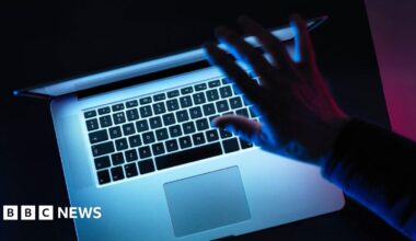 A bird eye view of a laptop in a dark room, with light coming from the screen. A hand hovers above the top of the laptop.