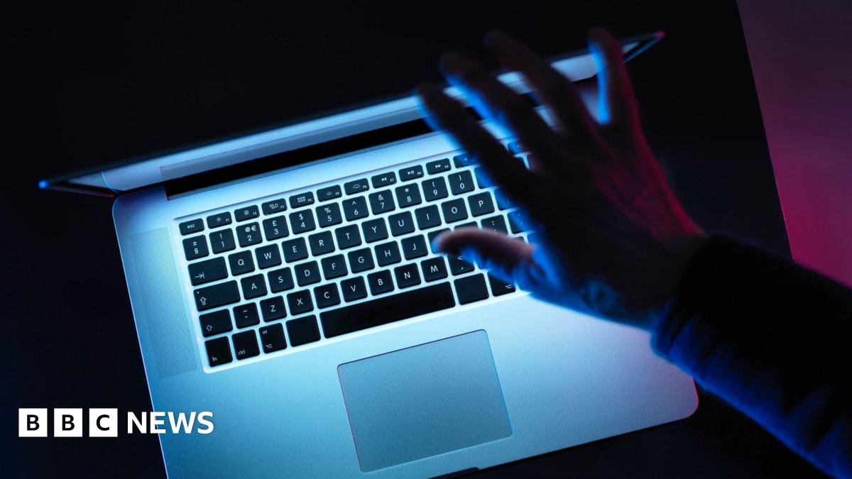 A bird eye view of a laptop in a dark room, with light coming from the screen. A hand hovers above the top of the laptop.