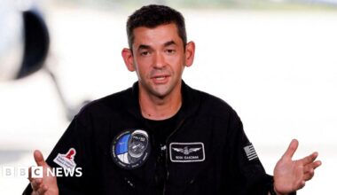 Jared Isaacman is wearing a black jumpsuit with Polaris patches on the chest. He has short brown hair and is gesturing with both hands as he speaks.