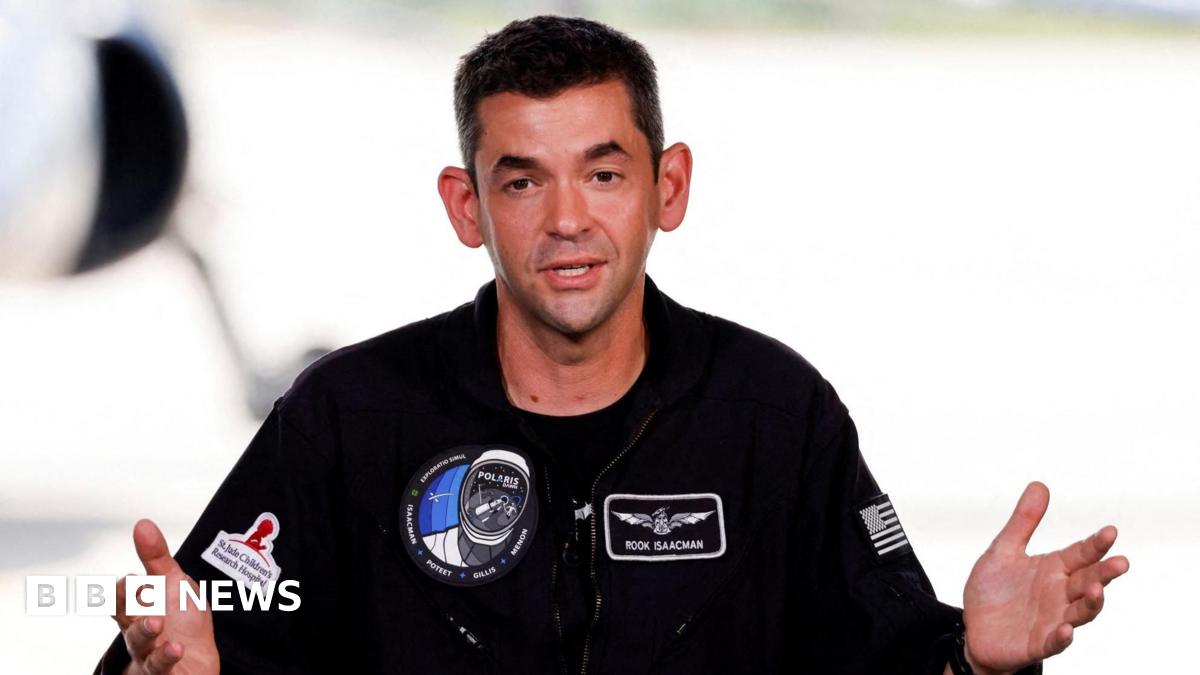 Jared Isaacman is wearing a black jumpsuit with Polaris patches on the chest. He has short brown hair and is gesturing with both hands as he speaks.