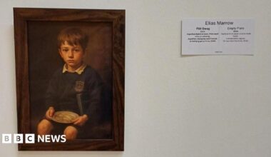 A brown frame that has an image of a school boy holding a round plate on a white wall. There is a white piece of paper next to it that has Elias Marrow and Empty Plate 2024 written on it.