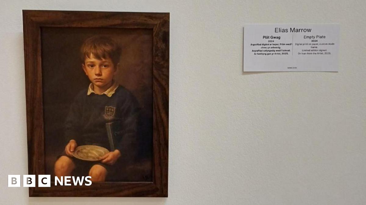 A brown frame that has an image of a school boy holding a round plate on a white wall. There is a white piece of paper next to it that has Elias Marrow and Empty Plate 2024 written on it.
