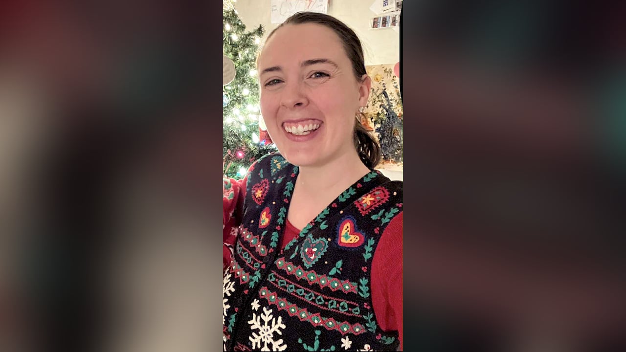 A woman in a festive vest smiles in front of a Christmas tree