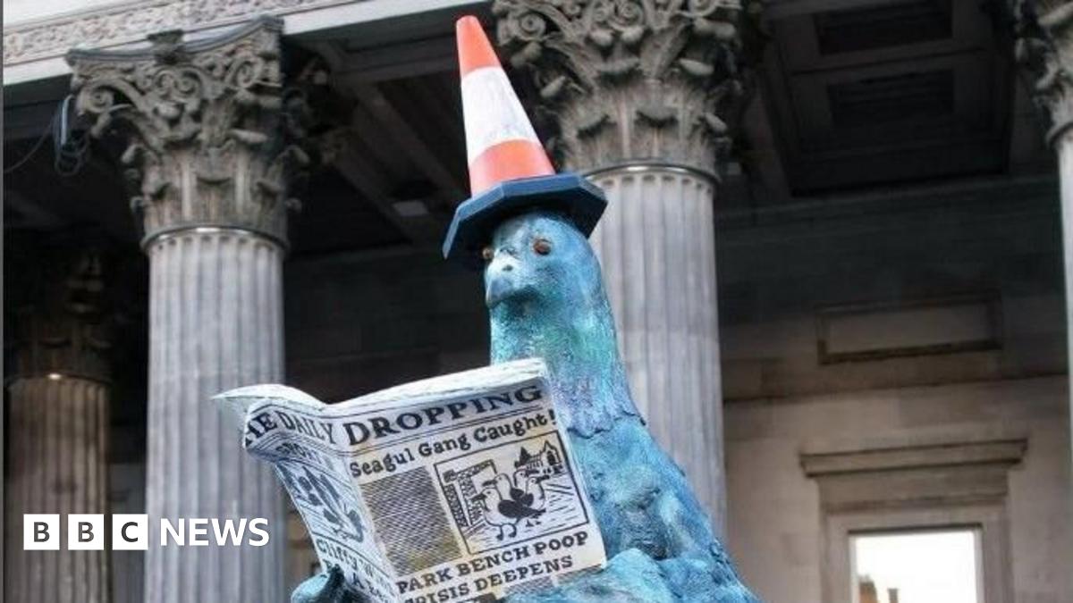 A view from behind of a statue, showing a pigeon with a traffic cone sitting on top of another statue.