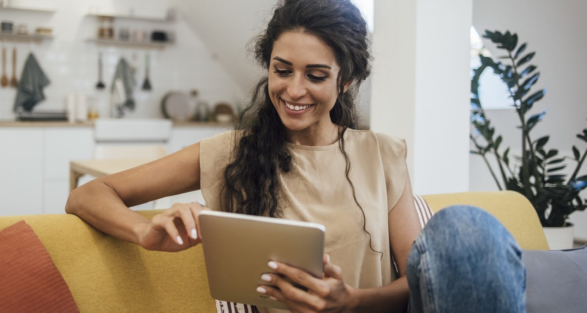 Happy young woman sitting on a couch outside of her kitchen and using a tablet next to a plant in her home