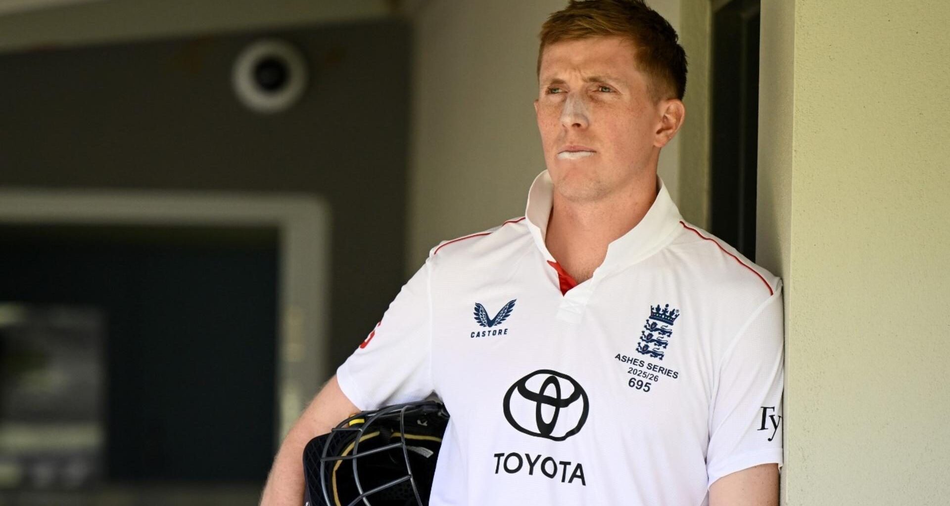 Zak Crawley leans against a wall holding his helmet under his arm
