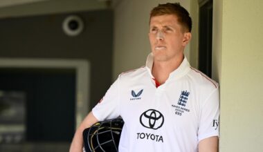 Zak Crawley leans against a wall holding his helmet under his arm