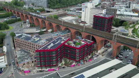 BBC A drone image of Stockport town centre. There is a train travelling along the historic viaduct which spans the length of the picture. In the middle of a ring road is a number of new, modern high rises located next to former mill buildings.