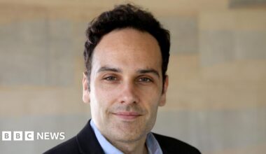 Dr Javier Andreu-Perez is smiling. He has black hair and stubble. He is wearing a black suit jacket and a pale blue shirt. There is a wall behind him, although it has been blurred.