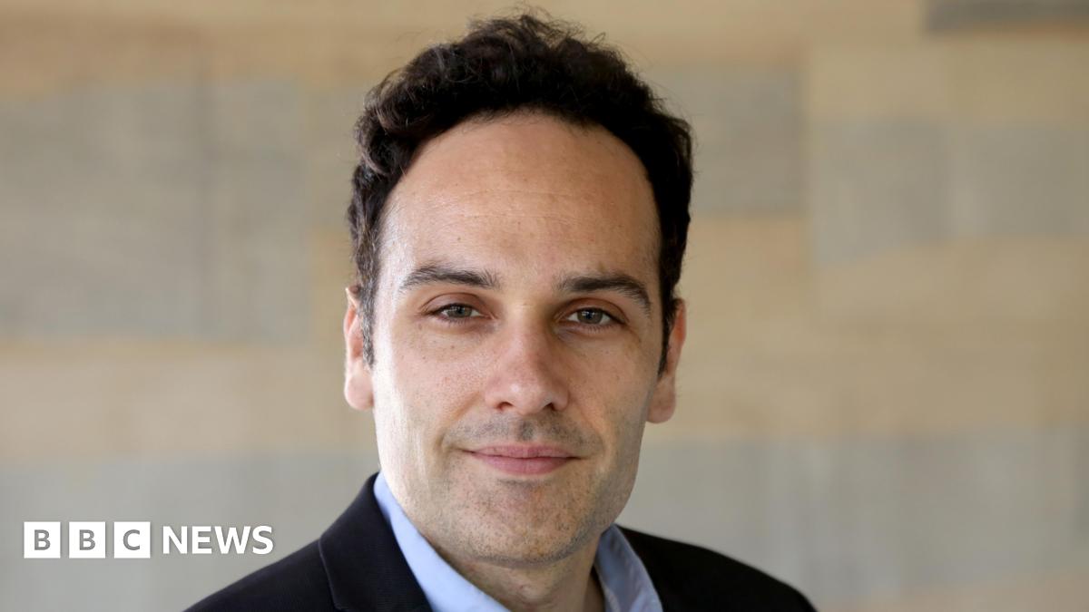 Dr Javier Andreu-Perez is smiling. He has black hair and stubble. He is wearing a black suit jacket and a pale blue shirt. There is a wall behind him, although it has been blurred.