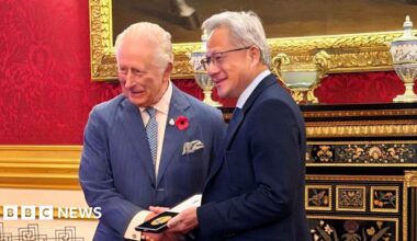 King Charles handing a prize to Jensen Huang - the King is wearing a blue pinstripe suit and has a poppy on his lapel, Jensen Huang is holding the gold medal in its case and is wearing a dark suit and smiling with glasses on - the room they are in is very grand, with gold lining around a big mirror behind them, and red wall paper