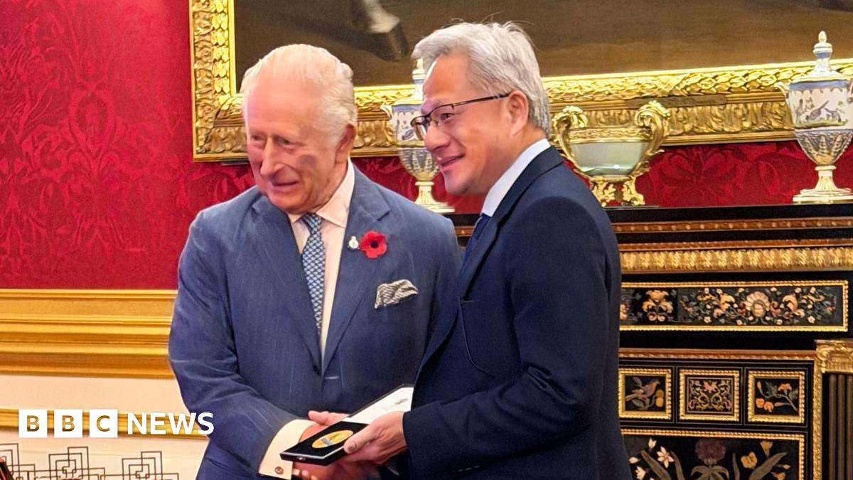 King Charles handing a prize to Jensen Huang - the King is wearing a blue pinstripe suit and has a poppy on his lapel, Jensen Huang is holding the gold medal in its case and is wearing a dark suit and smiling with glasses on - the room they are in is very grand, with gold lining around a big mirror behind them, and red wall paper