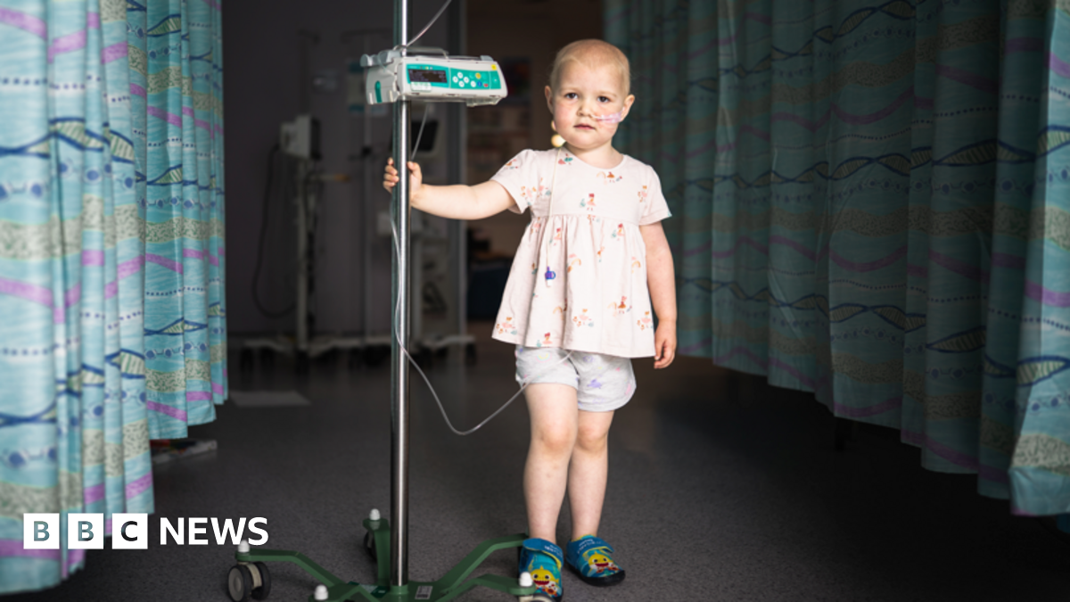 A photo of a little girl standing in the camera, she is wearing shorts and a top and holding onto a machine.