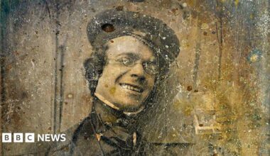 A sepia image of photography pioneer George Shaw. He is wearing Victorian clothes including a cap, shirt, jacket and neck tie. He is wearing oval glasses and smiling.