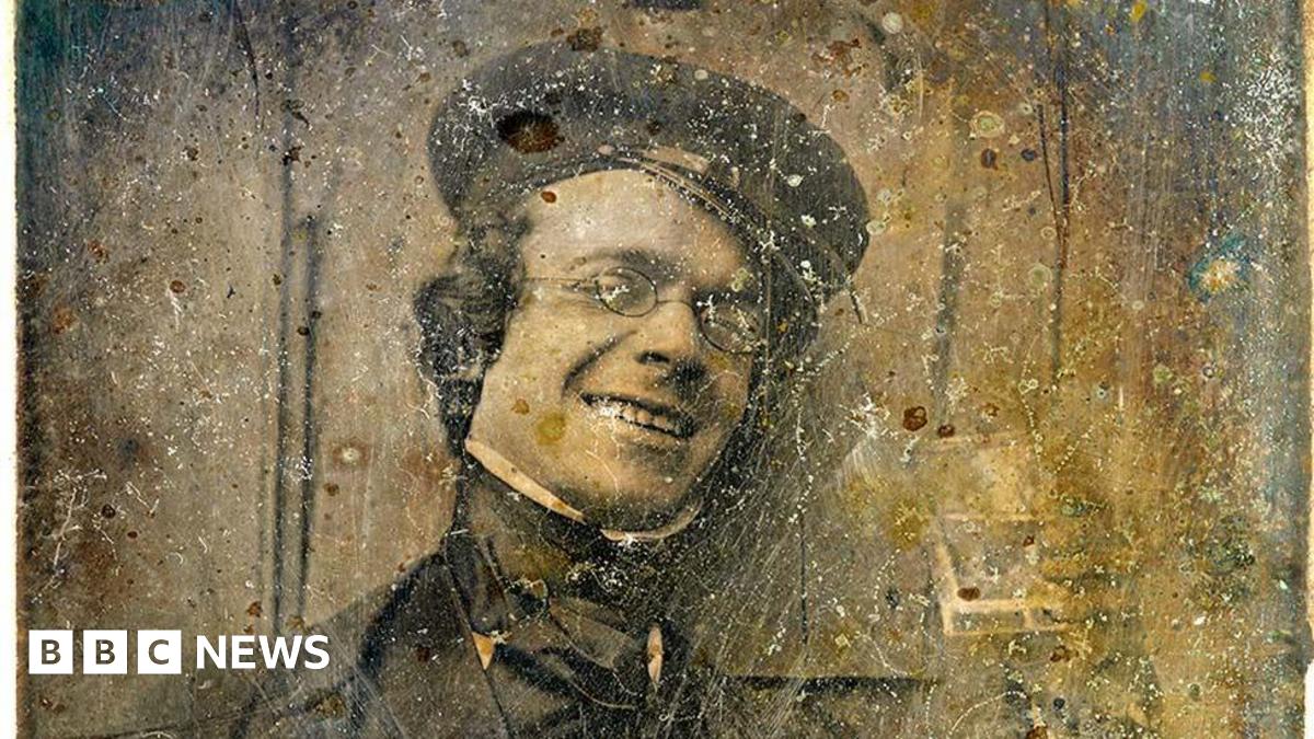 A sepia image of photography pioneer George Shaw. He is wearing Victorian clothes including a cap, shirt, jacket and neck tie. He is wearing oval glasses and smiling.