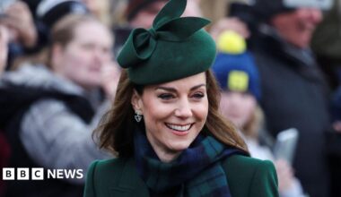 She is smiling and wearing dark green and a matching green hat. She has a green and navy checkered scarf around her neck.