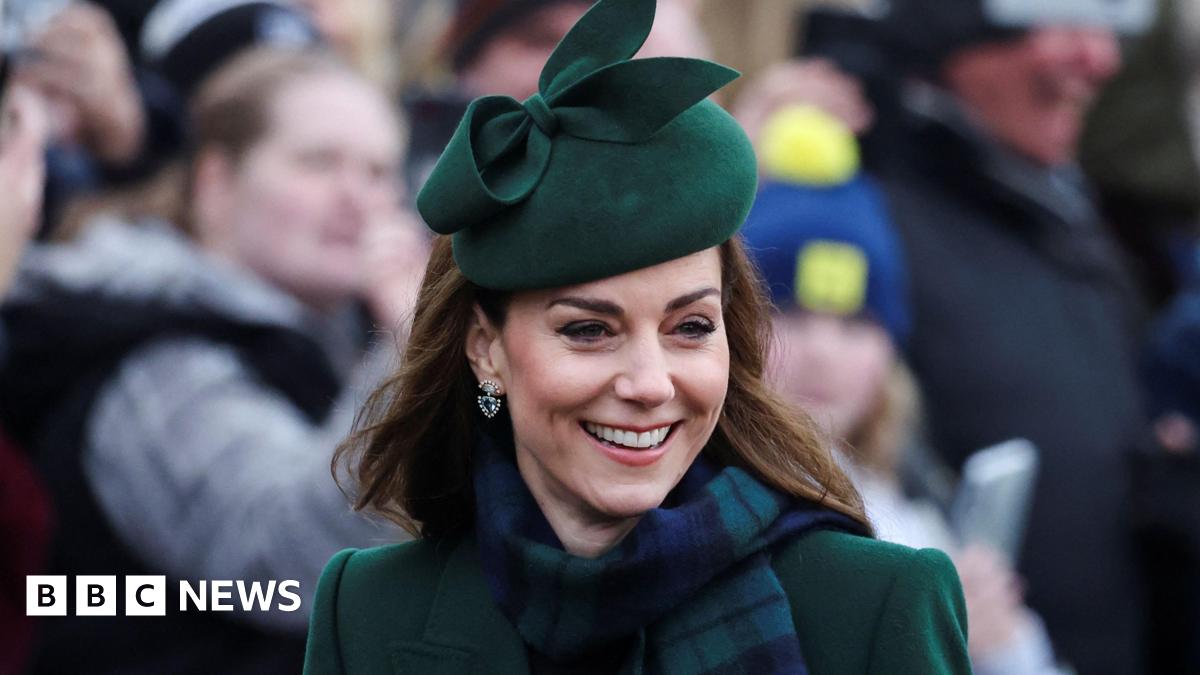 She is smiling and wearing dark green and a matching green hat. She has a green and navy checkered scarf around her neck.