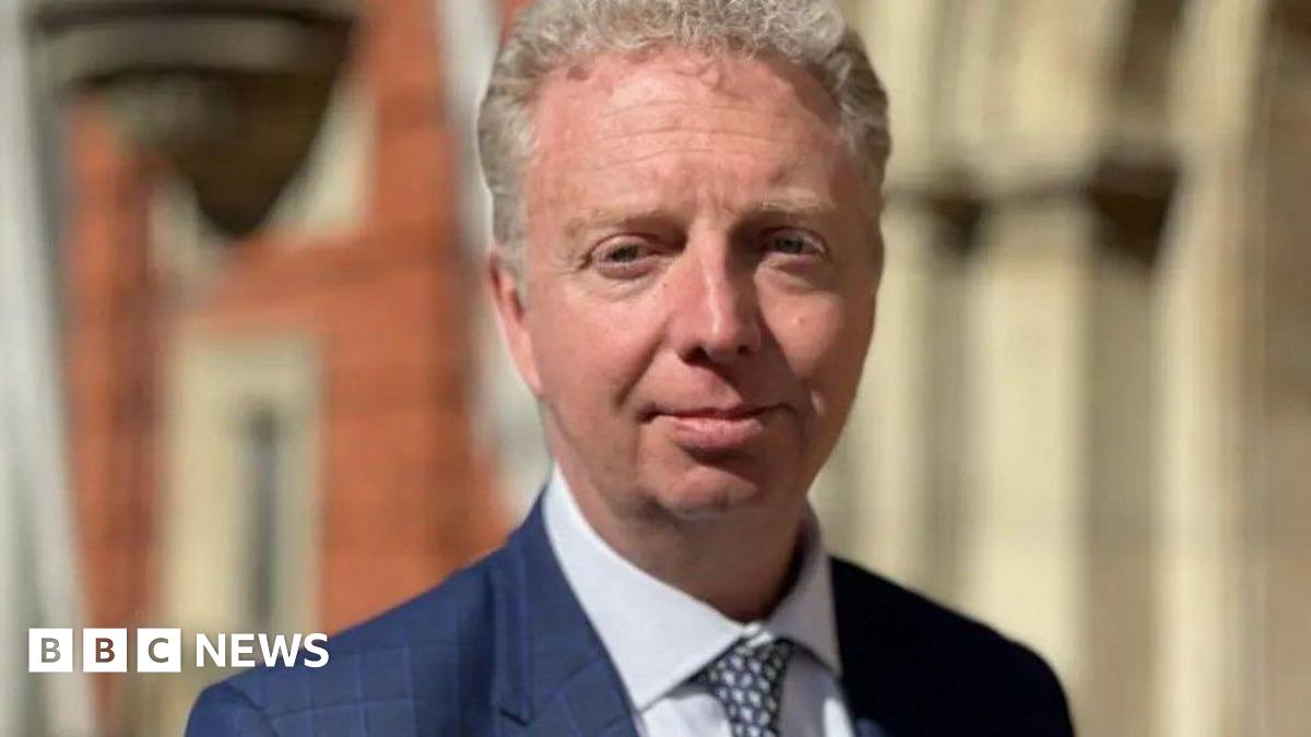Alex Allinson, who has short wavy light coloured hair and is wearing a blue jacket and tie with a white shirt.