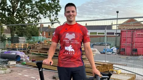 George Fox George Fox in a red technical T-shirt with 'George Fox Cycling Solutions" written on the chest. He is holding the saddle and handlebars of a black bike and standing against the backdrop of a construction site.