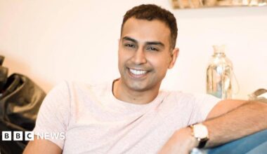 A man who appears to be in his 30s - he is sitting on a sofa at home, wearing a light off-white t-shirt, and smiling, clearly relaxed in the setting - he is leaning on the arm of the sofa with his left arm, and his watch is visible