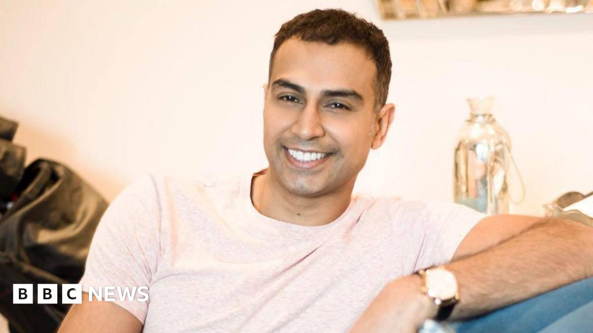 A man who appears to be in his 30s - he is sitting on a sofa at home, wearing a light off-white t-shirt, and smiling, clearly relaxed in the setting - he is leaning on the arm of the sofa with his left arm, and his watch is visible