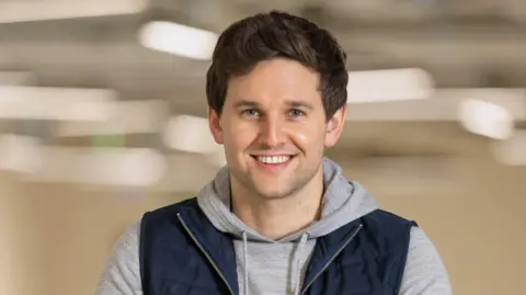 Net World Sports A man in his 30s smiling at the camera, he has brown hair and wears a gray hoodie and navy zip-up vest. The background is beige and blurred.