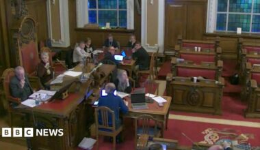 People in city hall chambers, with a red carpet and wooden benches. A few people are still seated around a table but there's nobody in the pulpits.