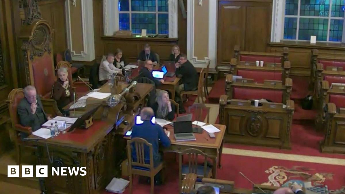 People in city hall chambers, with a red carpet and wooden benches. A few people are still seated around a table but there's nobody in the pulpits.