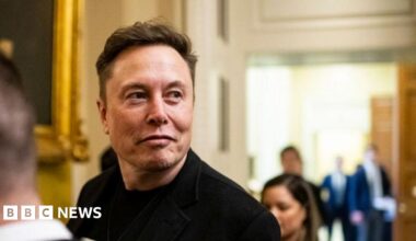 Elon Musk wearing a dark jacket. He is looking away from the camera with a smirk on his face