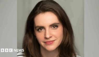 A close-up picture of Clementine Collett. She has long brown hair and brown eyes and is wearing a black and white striped T-shirt. Behind her is a grey background.