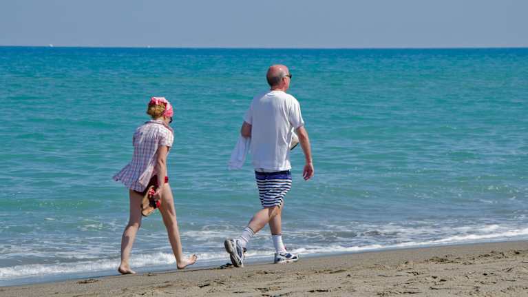A man and a woman with greying and thinning hair walk along a beach in swimwear, with an expanse of blue ocean seen in the background.