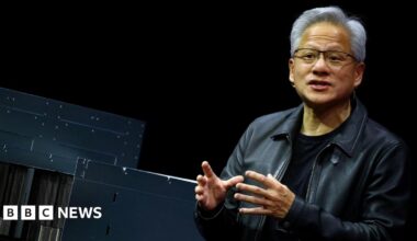 A man wearing a leather jacket speaks with his hands raised. A logo reading "Nvidia" is seen in the background.