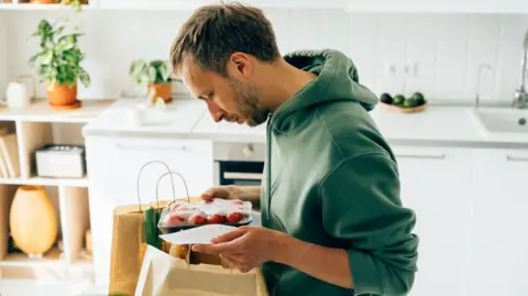 Getty Images Man stands in his kitchen with two shopping bags and holds tomatoes and a receipt in his hands
