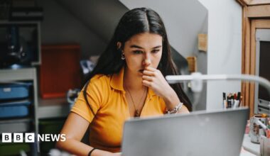 A young woman in an orange top looks at her laptop while chewing her finger