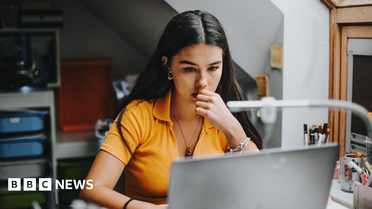 A young woman in an orange top looks at her laptop while chewing her finger