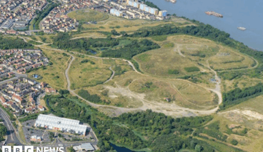 The river Thames can be seen on the right from an aerial shot - with green fields and trees, on the outskirts of which are low and high-rise housing and buildings with a lake in the foreground.