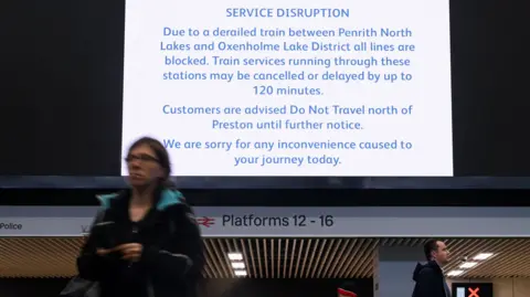 Reuters A sign at London Euston detailing the delay. There is a woman and a man walking underneath the sign. It reads 'SERVICE DISRUPTION- Due to a derailed train between Penrith North Lakes and Oxenholme Lake District all lines are blocked. Train services running through these stations may be cancelled or delayed by up to 120 minutes. Customers are advised Do Not Travel north of Preston until further notice. We are sorry for an inconvenience caused today.'