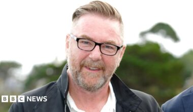Hollywood director Michael Caton Jones - a fair-haired man with a beard and glasses stands against a leafy background, wearing a black collared jacket.