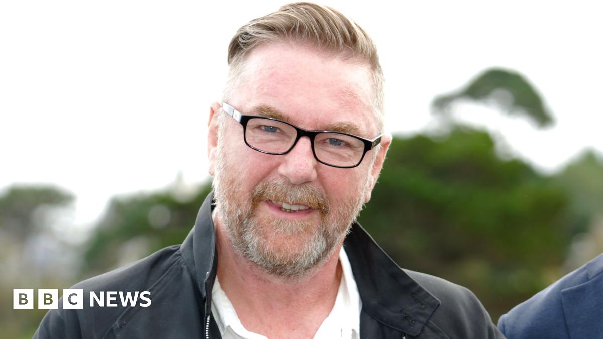 Hollywood director Michael Caton Jones - a fair-haired man with a beard and glasses stands against a leafy background, wearing a black collared jacket.