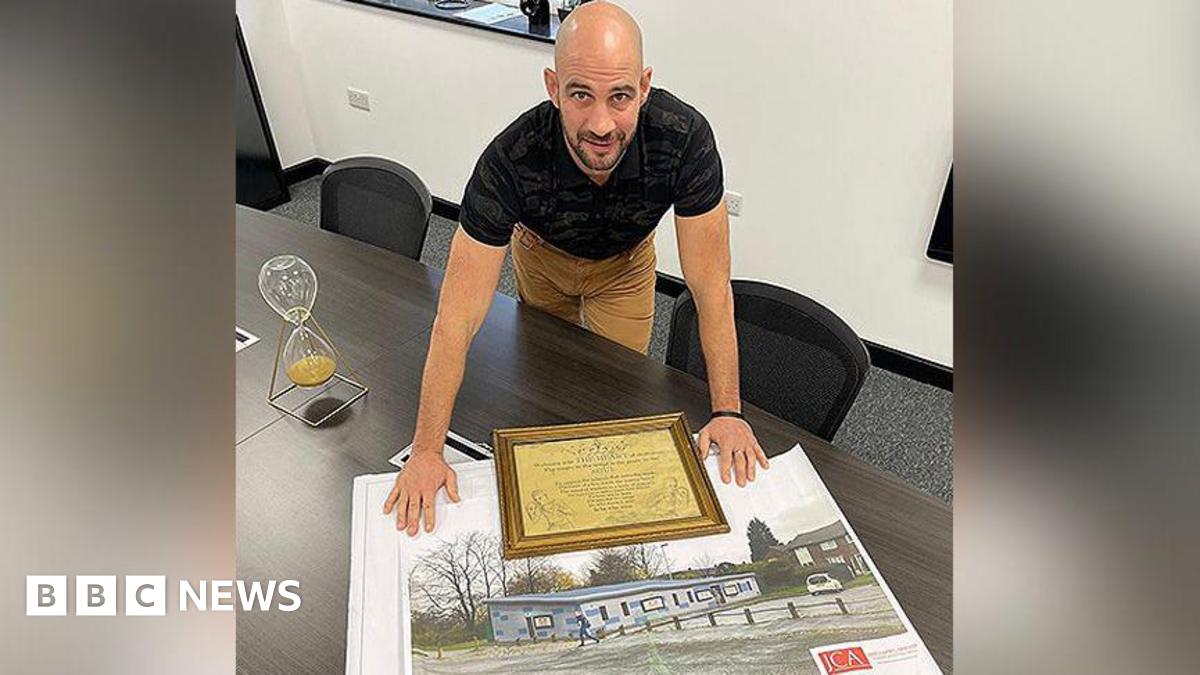 Dale Gaucas, wearing a black top and beige trousers, is leaning over a large photograph of the boxing club built in his old Darnhill estate.