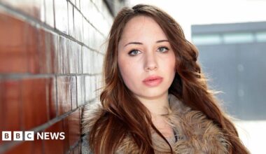 A close-up of Alice Figueiredo. She has long auburn hair and is looking directly at the camera. She is wearing a winter coat and is leaning against a brick wall.