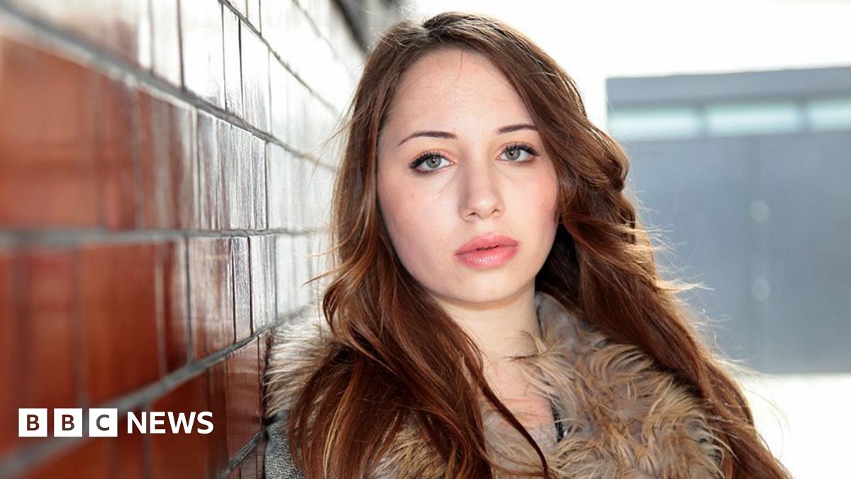 A close-up of Alice Figueiredo. She has long auburn hair and is looking directly at the camera. She is wearing a winter coat and is leaning against a brick wall.