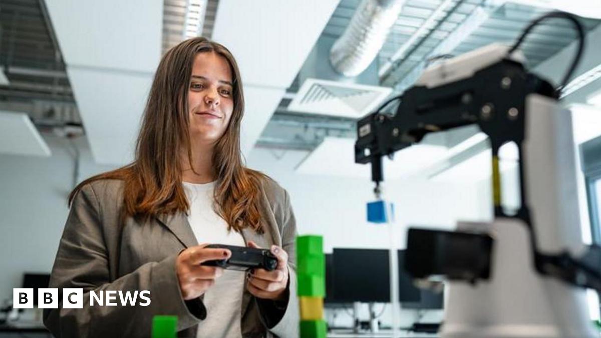 A woman with long brown hair, a white top and brown jacket holding a small black device in a white room with mechanical devices in it